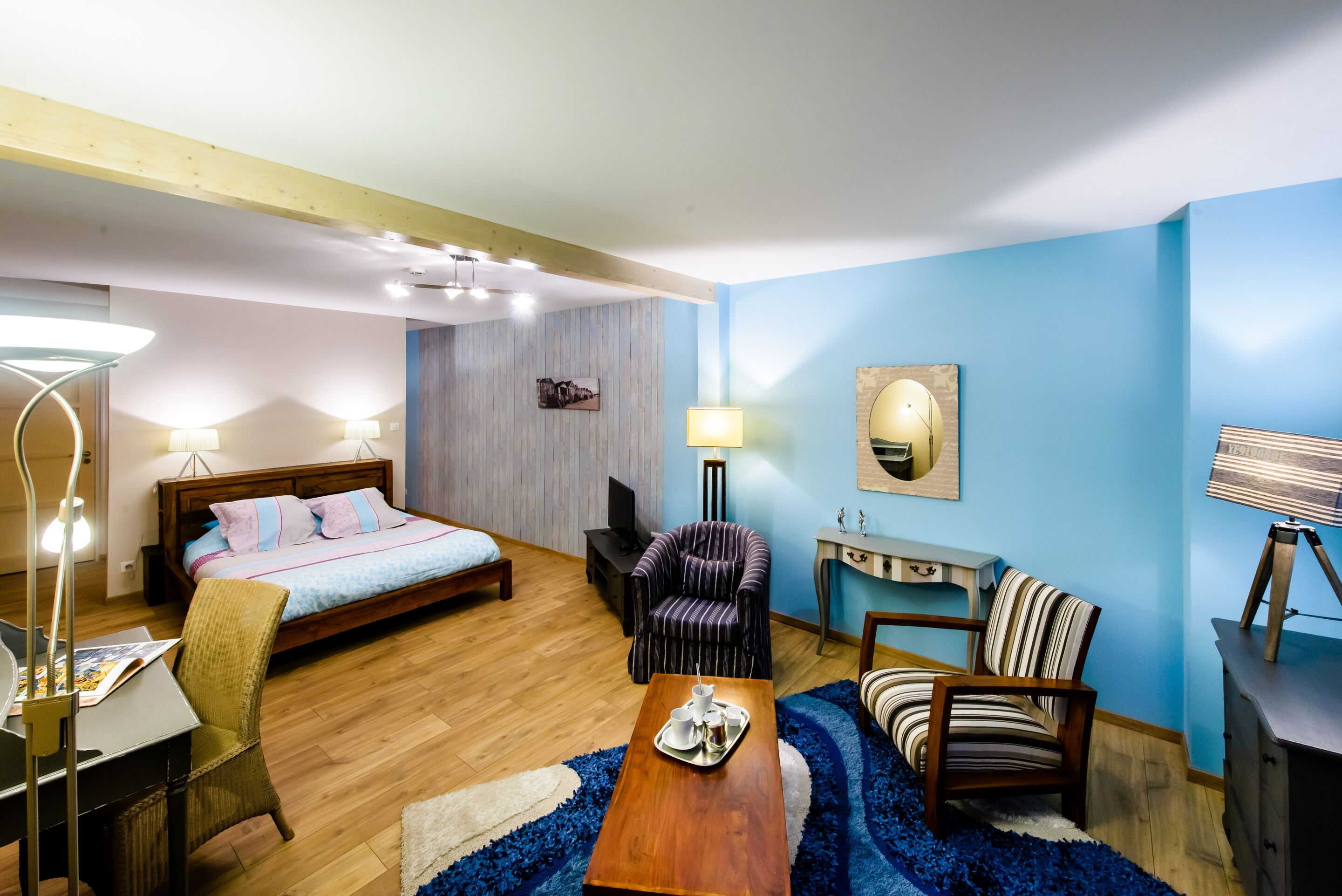 A hotel room with a bed, a desk with a chair, a small table with a tray, a chair, a lamp, and a wall-mounted TV.