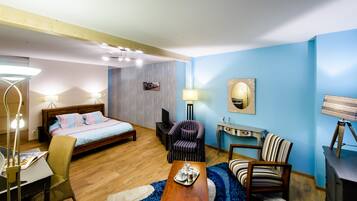 A hotel room with a bed, a desk with a chair, a small table with a tray, a chair, a lamp, and a wall-mounted TV.