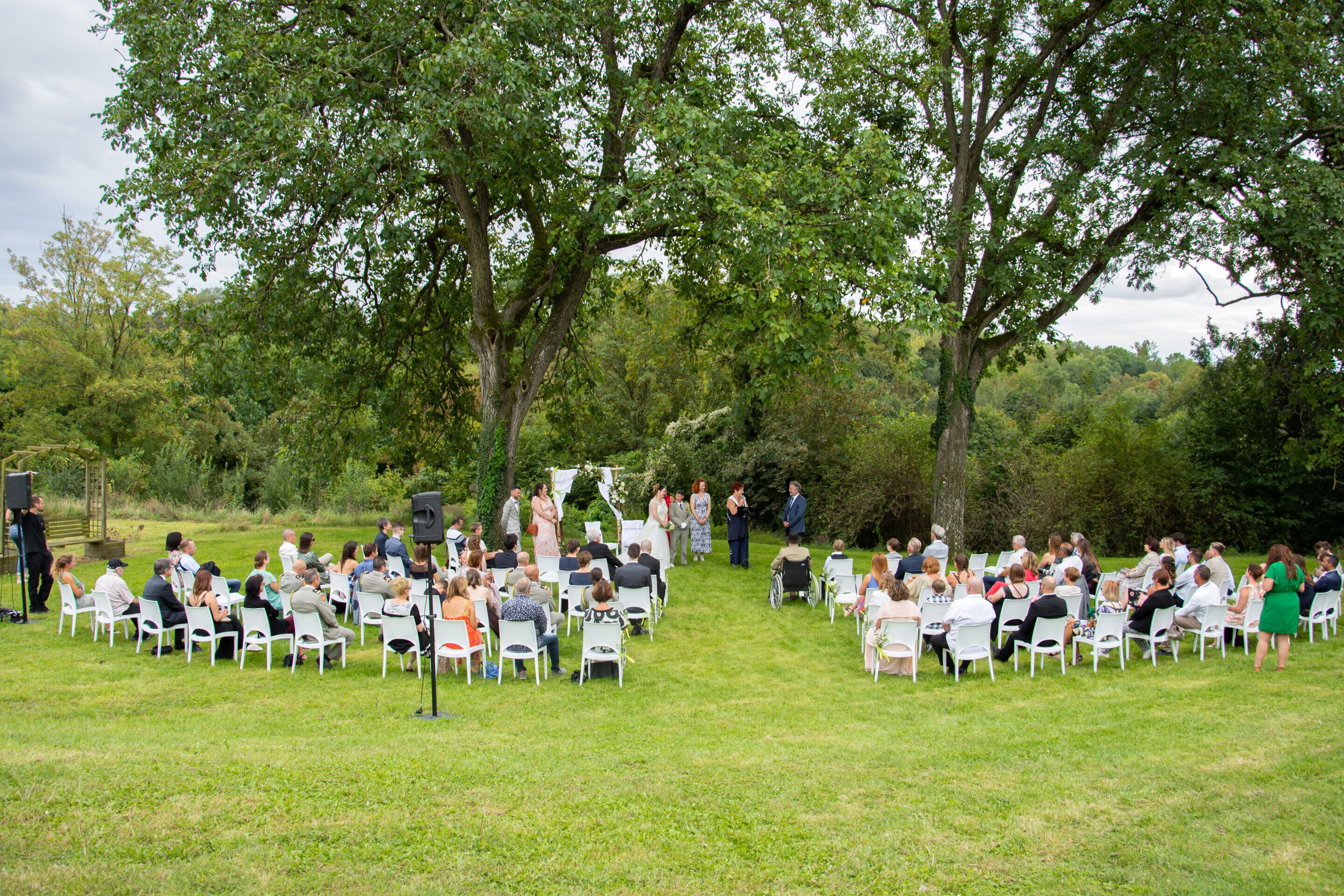 Een evenement buiten met rijen witte stoelen die in een halve cirkel zijn gerangschikt, omringd door bomen.