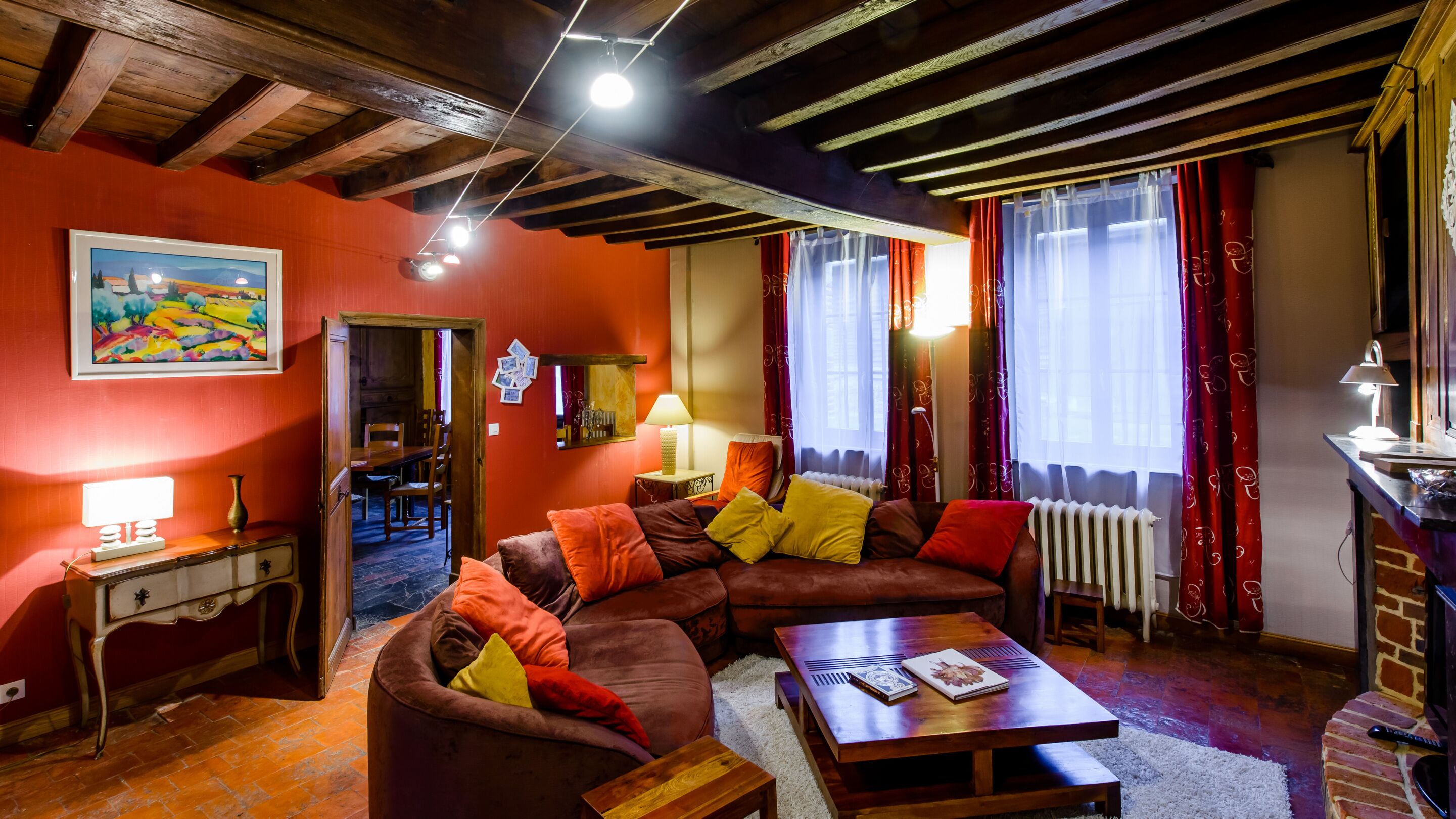 Ein gemütliches Wohnzimmer mit Holzbalkendecke, roten Wänden, einer Sofagarnitur, einem Couchtisch und einem Essbereich, der durch einen offenen Durchgang sichtbar ist.