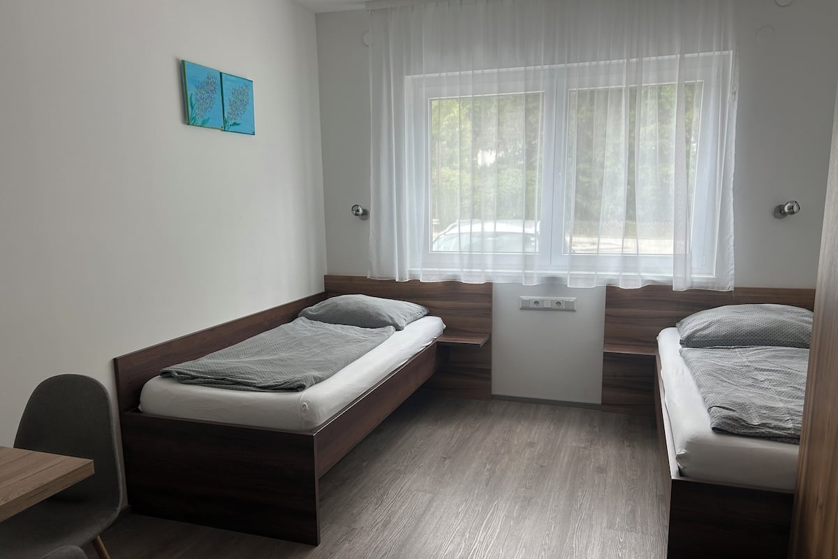 Une petite chambre moderne avec deux lits simples, un bureau et une fenêtre ornée de rideaux légers.