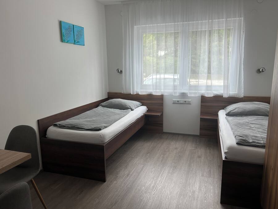 A small, modern room with two single beds, a desk, and a window with sheer curtains.