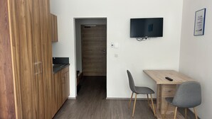 A compact room with a kitchenette, a bed, a desk with a chair, a TV, and a door leading to another room.
