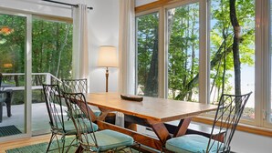 Een eethoek met een houten tafel en stoelen, grote ramen met uitzicht op bomen, en een lamp op tafel.