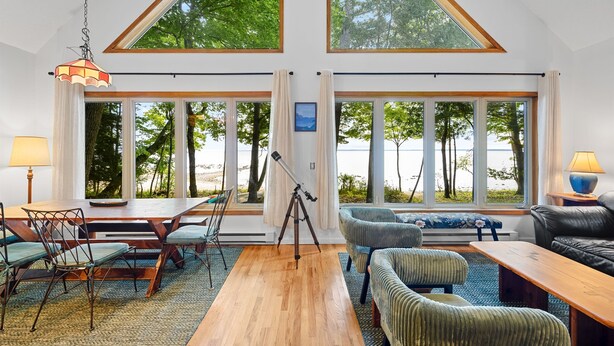 Een ruime woonkamer met een houten vloer, een eethoek met een tafel en stoelen, een statief en grote ramen met uitzicht op bomen en water.