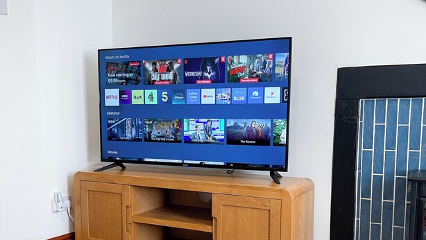 Uma sala de estar com televisão de tela plana exibindo a interface de um serviço de streaming, um rack de madeira, uma lareira e uma revista sobre a mesa.