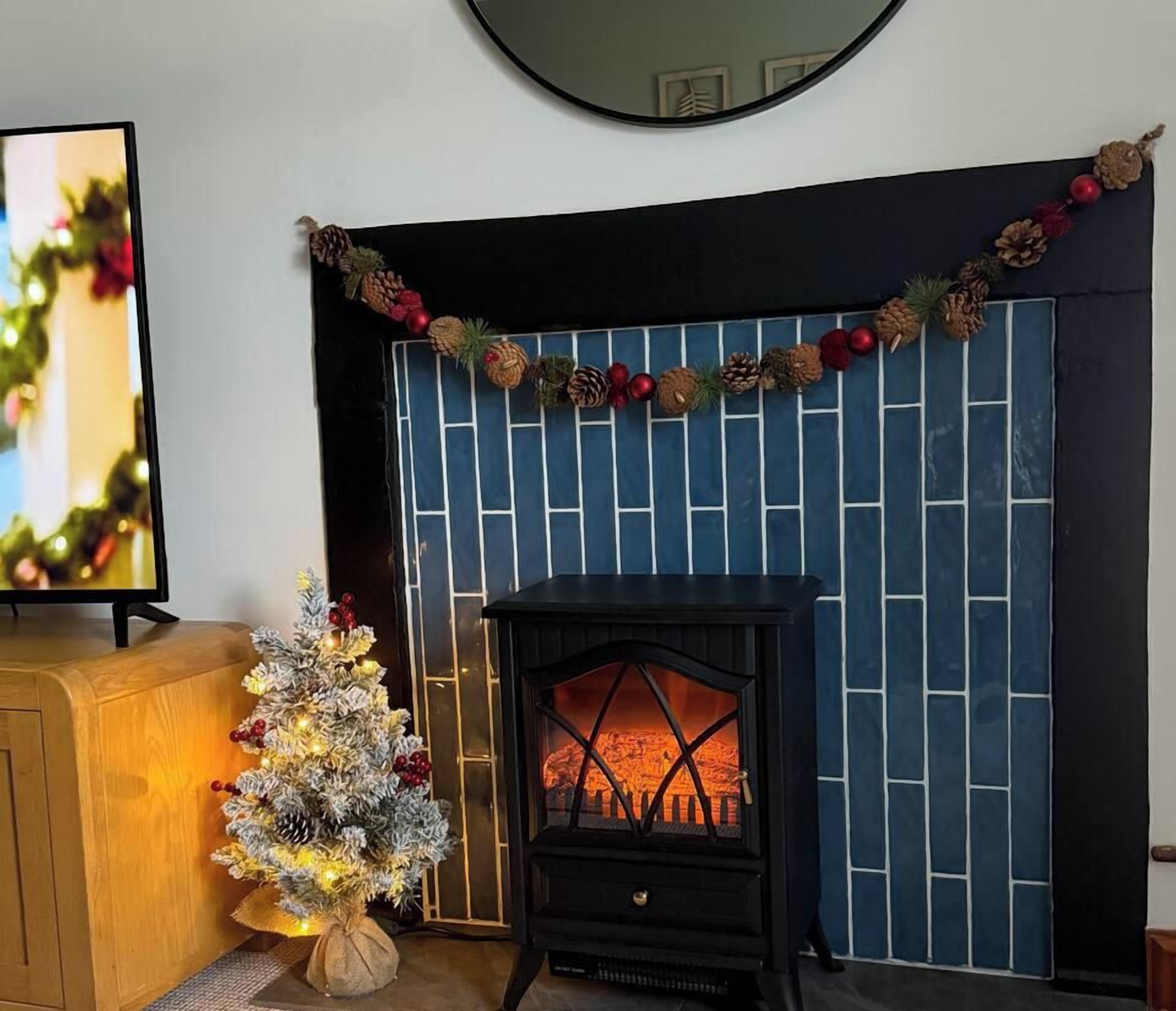 Ein Kamin mit dekorativer Girlande, eine kleine Weihnachtstanne mit Lichtern und ein Fernseher auf einem Holzschrank.