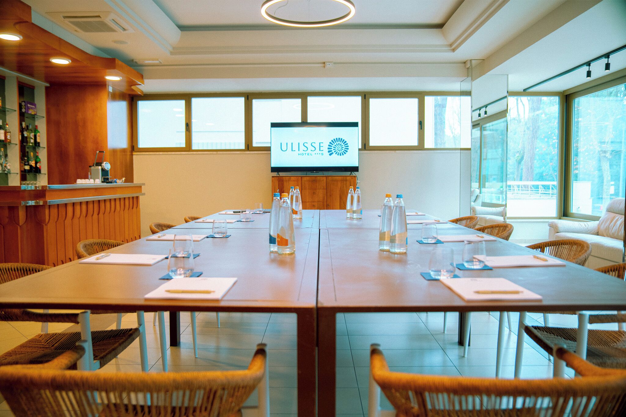 A conference room with a long table, chairs, a TV screen displaying the hotel name, and a bar area with bottles.