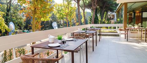 An outdoor patio with tables and wicker chairs, surrounded by trees and a view of a building.