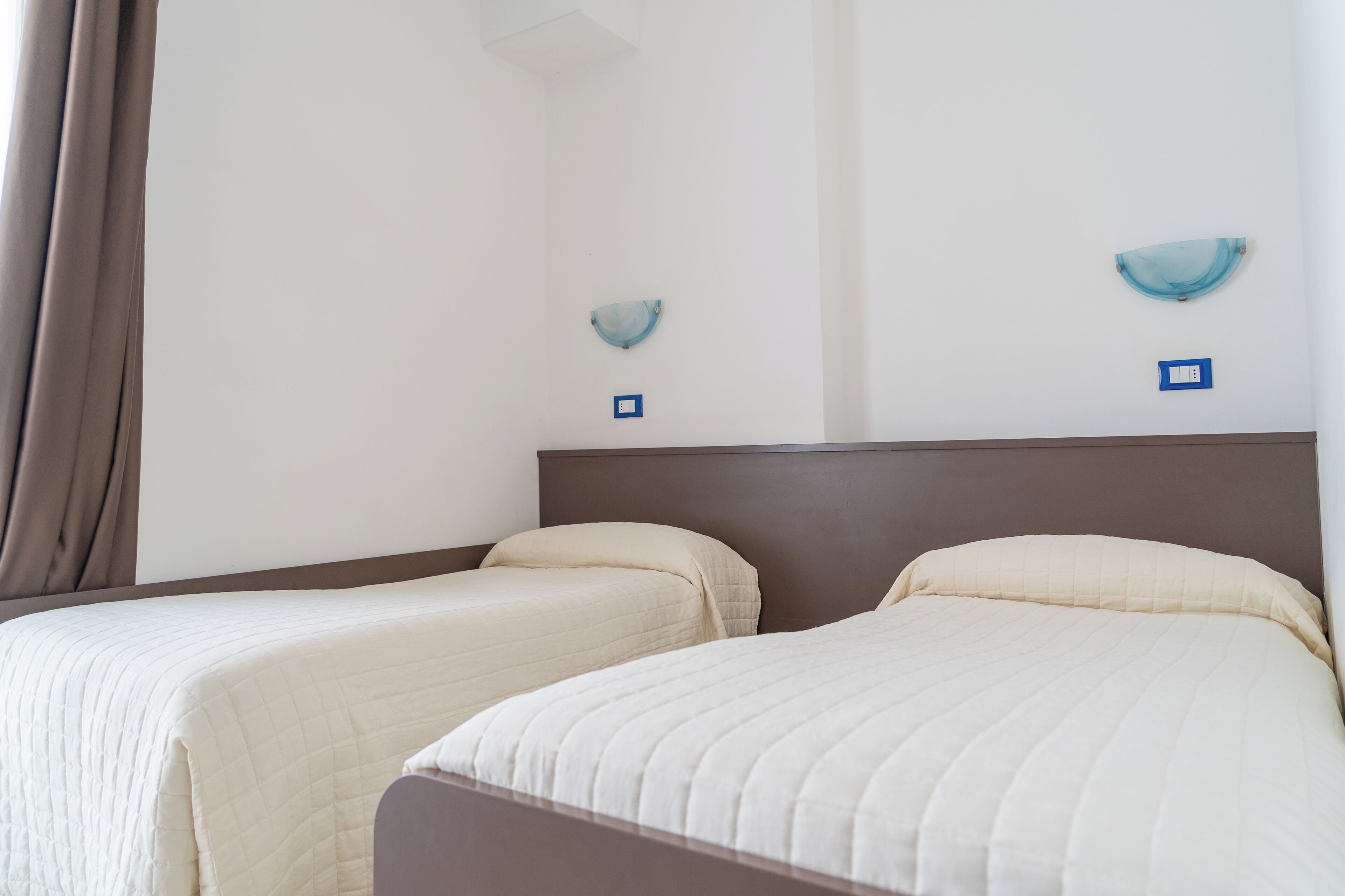 Camera d'albergo con due letti, una testiera scura e una parete di colore chiaro.