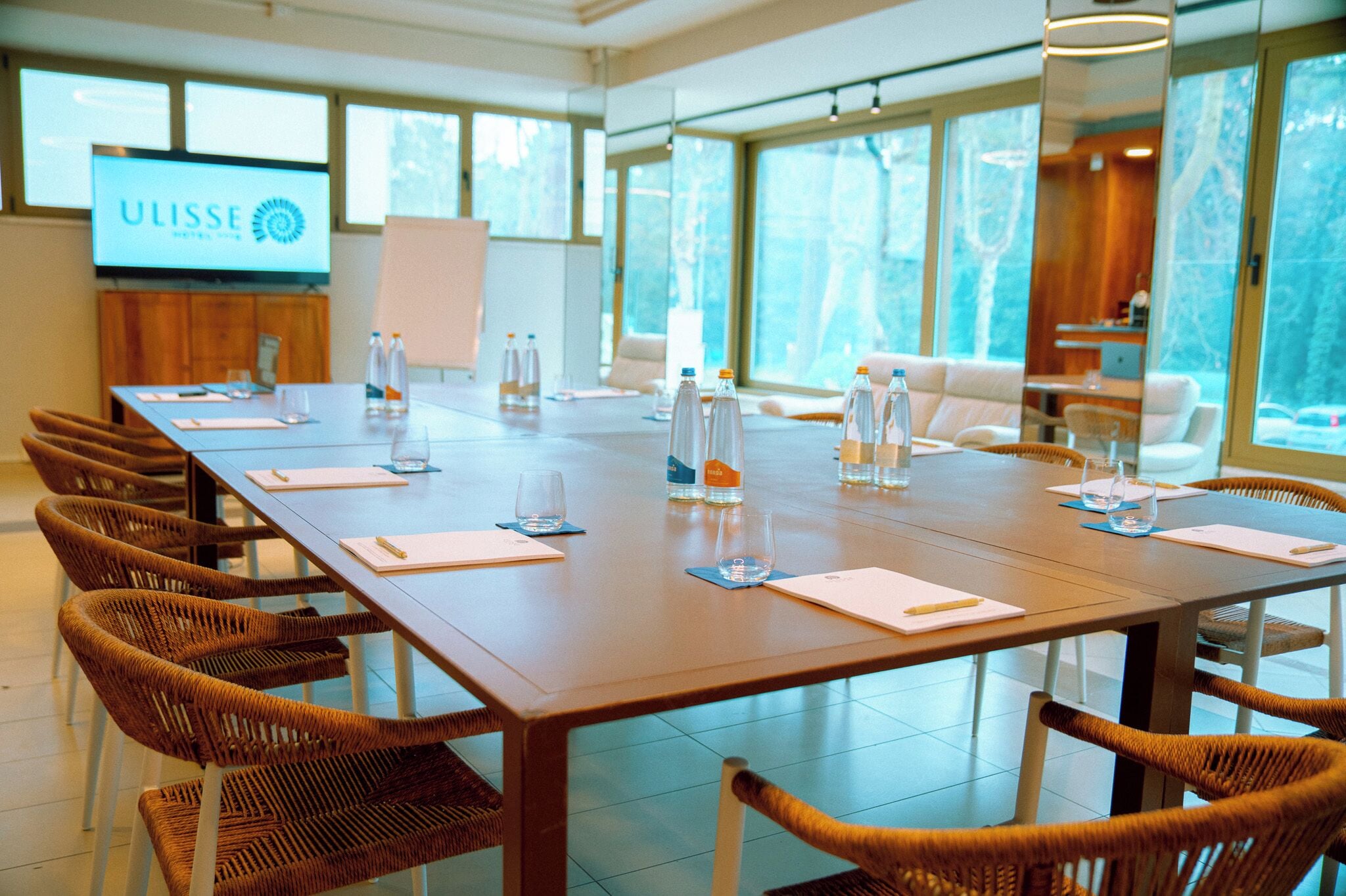 A conference room with a long table, wicker chairs, and a TV screen displaying 'ULISSE HOTEL'.