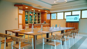 Een congreszaal met een lange tafel, rieten stoelen, een barruimte met flessenkasten en een televisiescherm waarop de naam van het hotel staat.