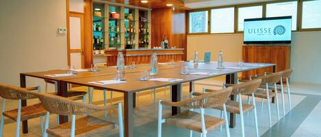 Un salón de conferencias con una larga mesa, sillas de mimbre, una zona de bar con estantes llenos de botellas y una pantalla de televisión mostrando el nombre del hotel.