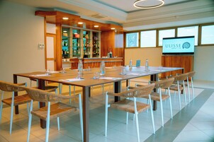 Un salón de conferencias con una larga mesa, sillas de mimbre, una zona de bar con estantes llenos de botellas y una pantalla de televisión mostrando el nombre del hotel.