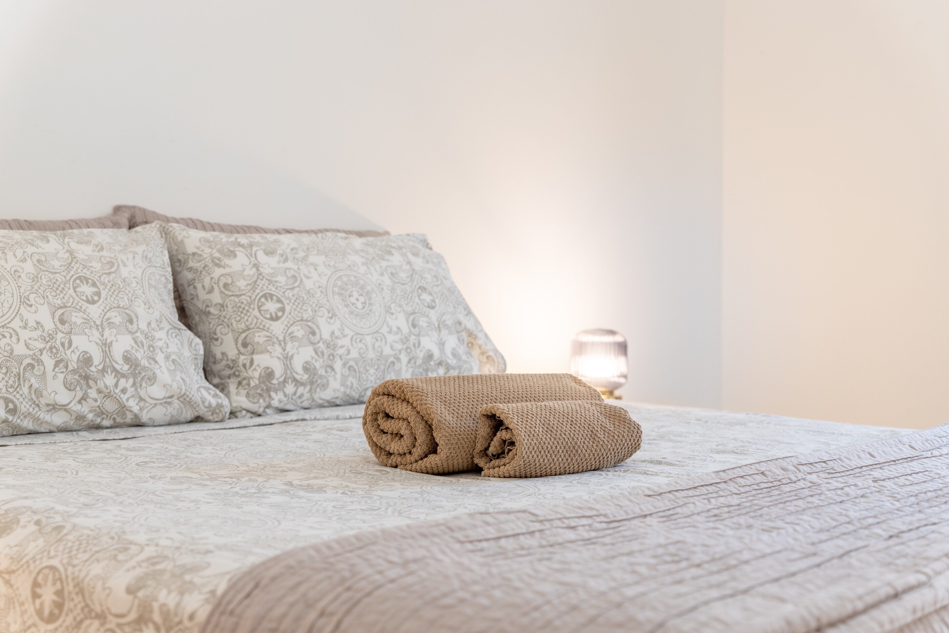 A neatly made bed with rolled towels, patterned pillows, and a bedside lamp.