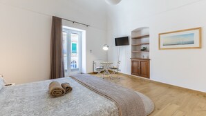A hotel room with a bed, a desk, a chair, a TV, a painting, and a view through a door. - Vico21 Apartment in Central Modica with Balcony, Wi-Fi, and Air Conditioning (Modica)