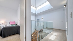 A modern bedroom with a skylight, a glass staircase, and a bed with a patterned bedspread.