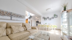 A modern living room with a beige sofa, a glass coffee table, and a dining area with a yellow and white tablecloth.