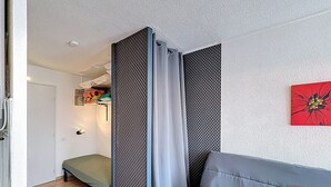 Ein Zimmer mit grauem Sofa, einem Bett, einer Trennwand mit strukturierter Musterung, einer Tür und einem Wandbild.