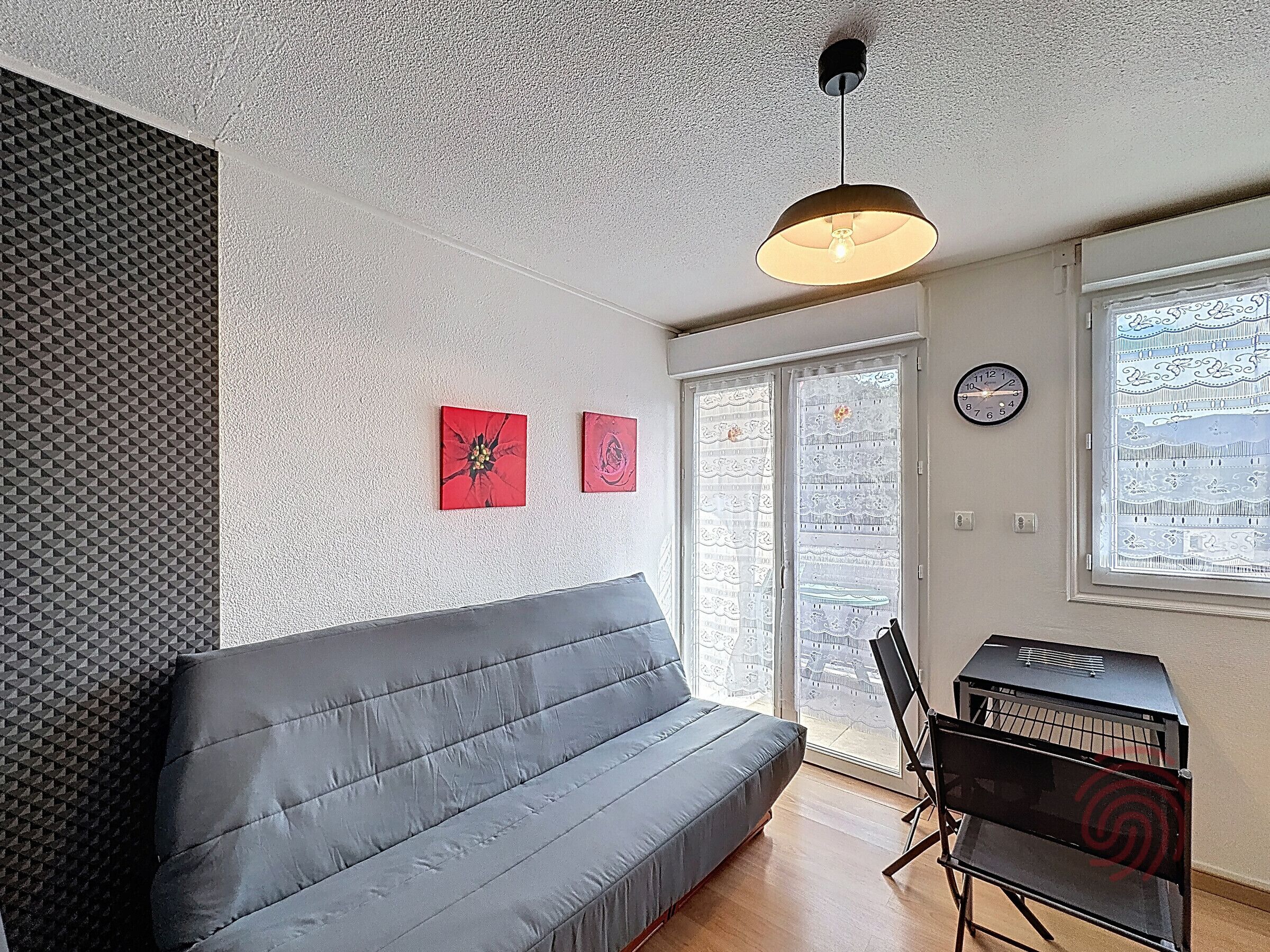 Un salon moderne avec un canapé gris, un luminaire au plafond, une horloge murale et une fenêtre donnant sur un beau paysage.
