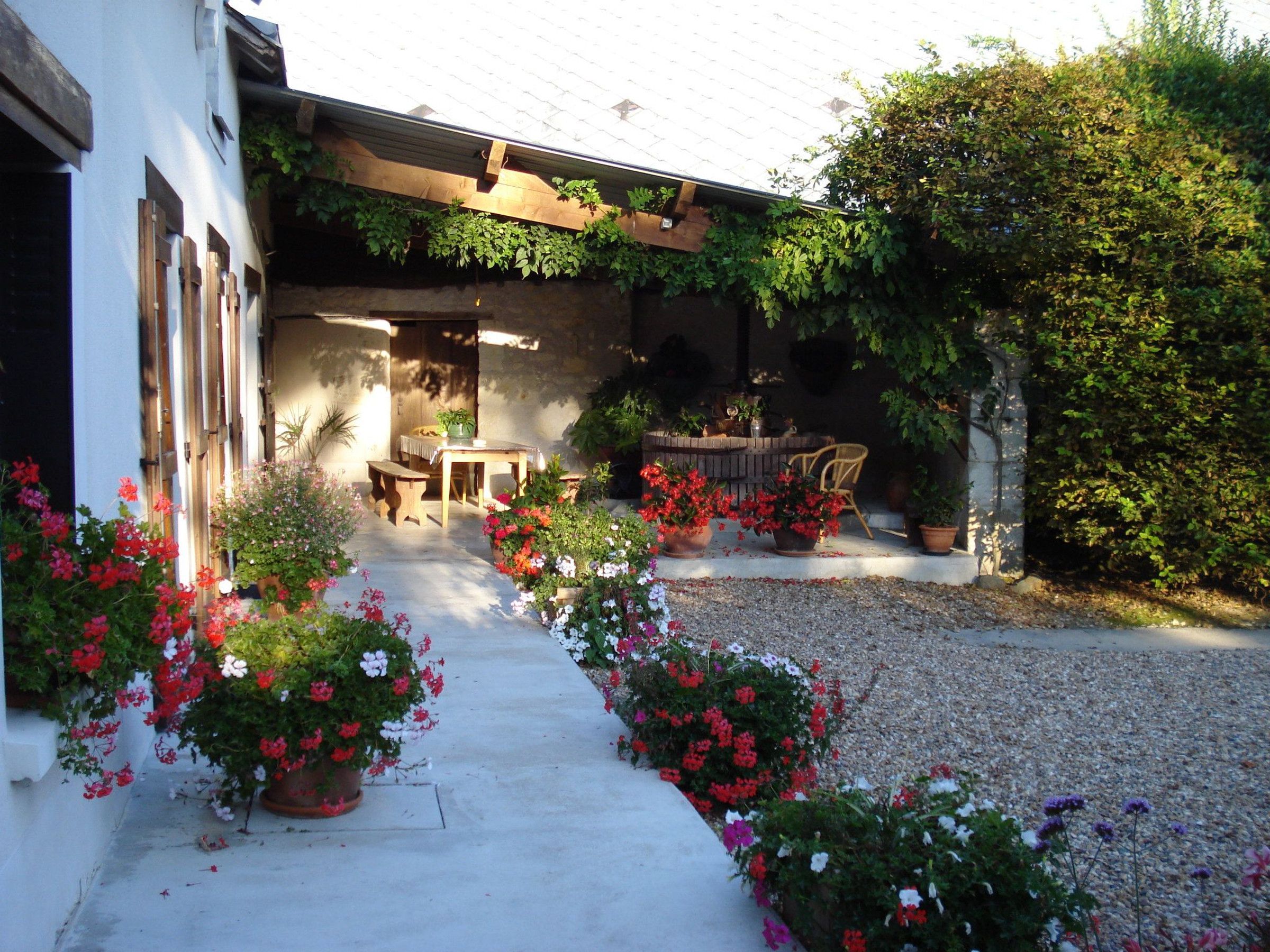 A courtyard with a wooden pergola, a table and chairs, and potted plants with flowers.