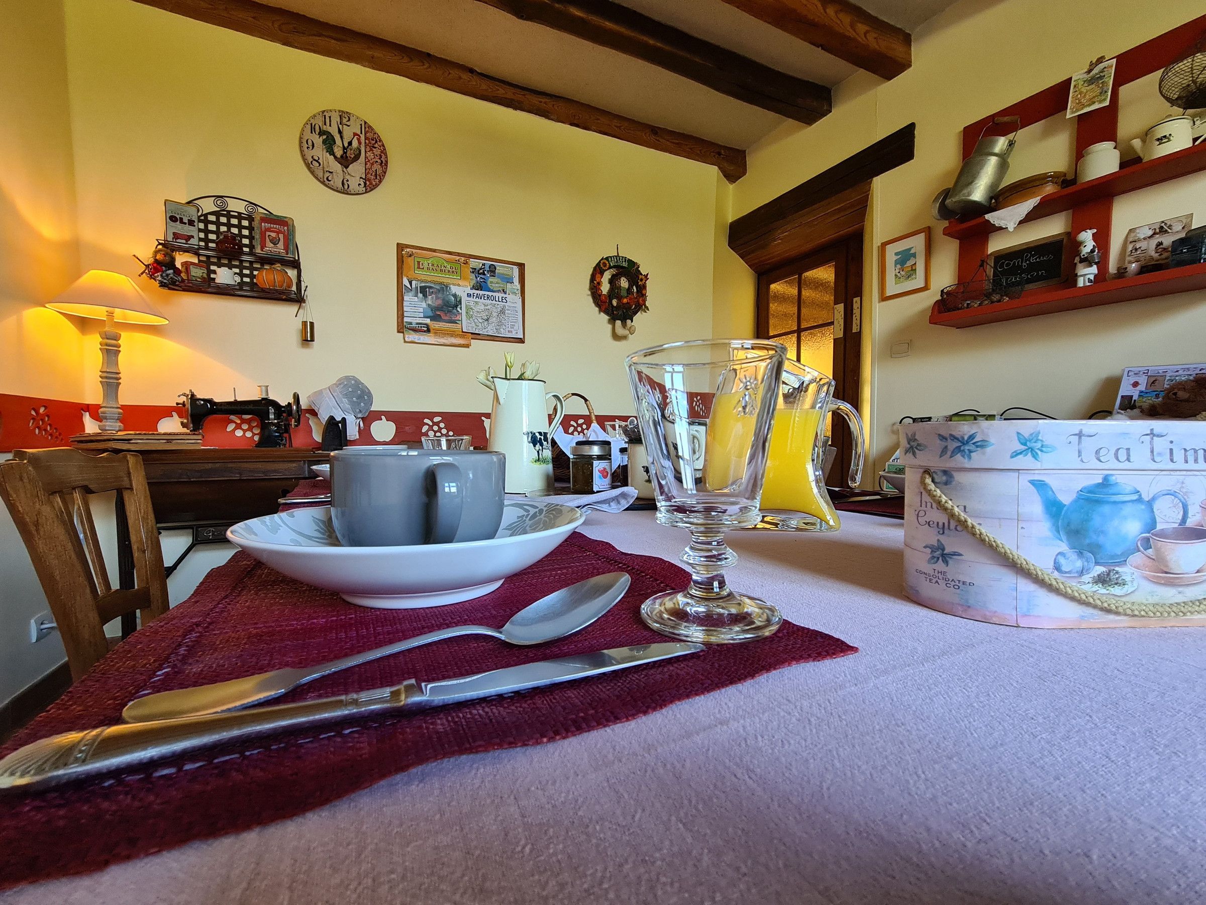 A dining table with a teapot, cups, and a jug, set in a cozy kitchen with wooden beams and a clock on the wall.