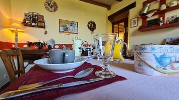 A dining table with a teapot, cups, and a jug, set in a cozy kitchen with wooden beams and a clock on the wall.