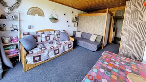 A cozy room with a wooden ceiling, a patterned sofa, a small table with a colorful tablecloth, and a decorative wall with a leaf pattern.