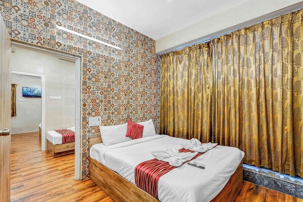 A hotel room with a patterned wall, a bed with white bedding and red accents, a wooden floor, and a window with curtains.