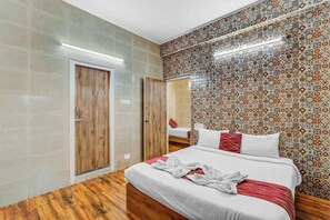A hotel room with a wooden floor, a bed with white bedding and red accents, a wooden wardrobe, and a patterned tile wall. - Season4 Residences 1-BHK furnished appt (Chennai)