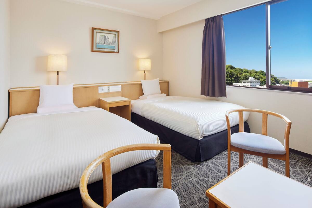 A hotel room with two beds, bedside tables, lamps, a chair, and a view of the city.