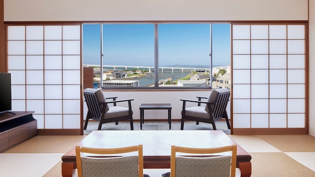 A room with tatami flooring, a low table, two chairs, and a large window with a view of a bridge and water.