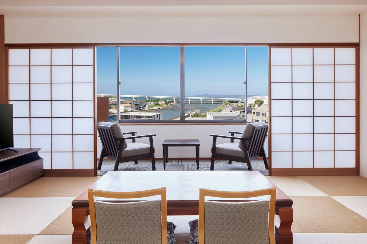 A room with tatami flooring, a low table, two chairs, and a large window with a view of a bridge and water.