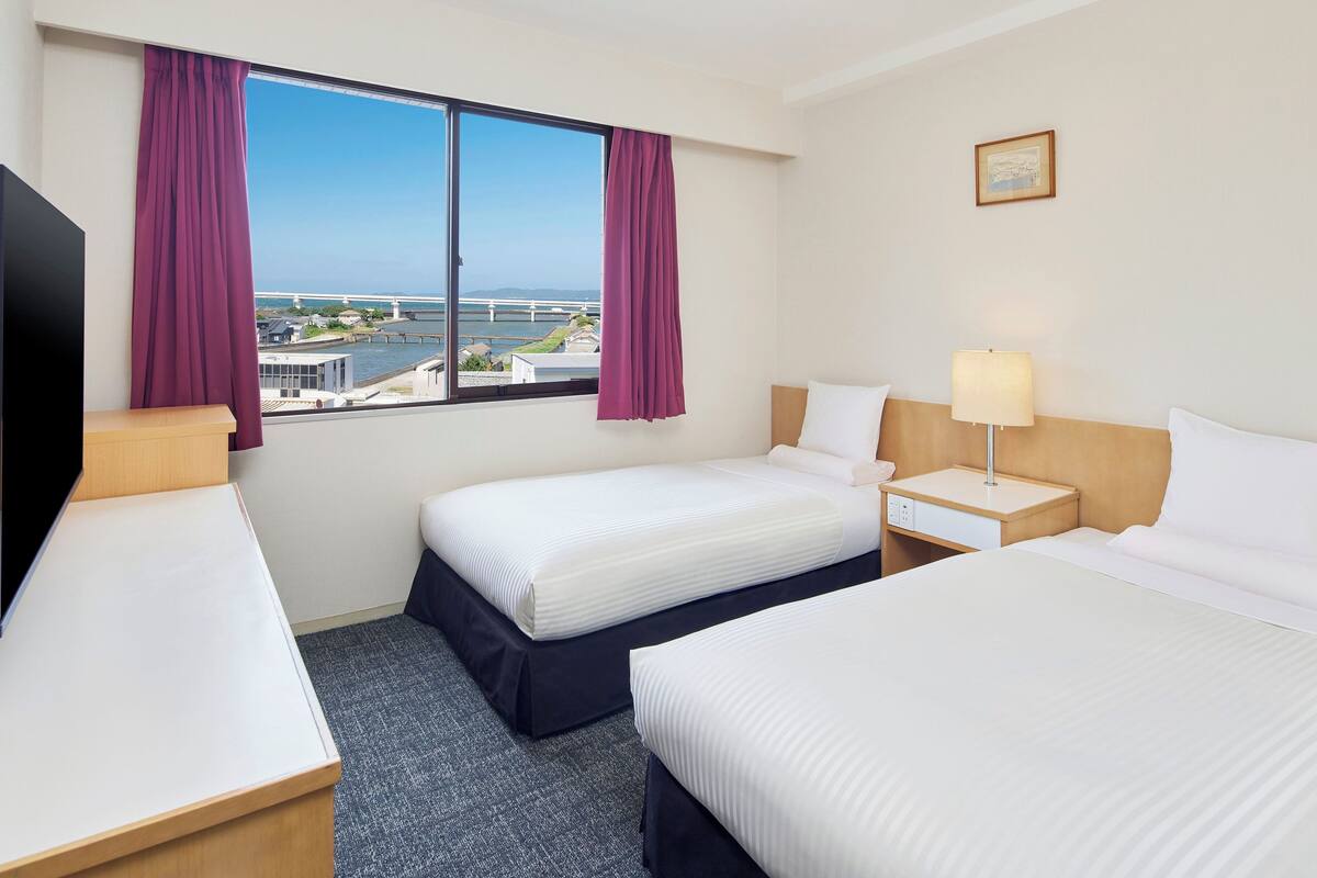 A hotel room with two beds, a desk, a chair, a lamp, and a window with curtains.