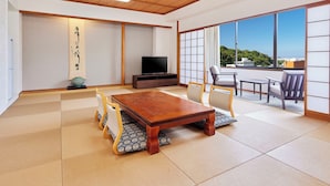 A traditional Japanese-style room with tatami flooring, a wooden ceiling, a television, and a sliding door leading to a balcony with a view.
