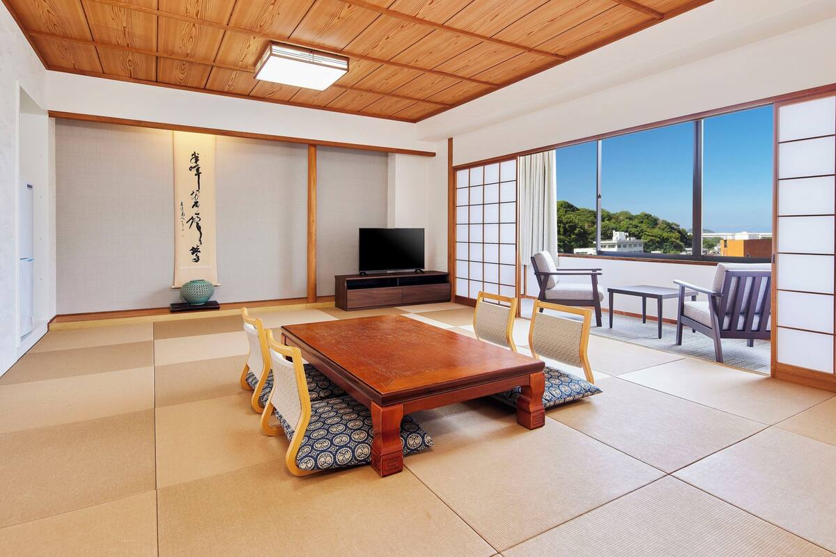 A traditional Japanese-style room with tatami flooring, a wooden ceiling, a television, and a sliding door leading to a balcony with a view.