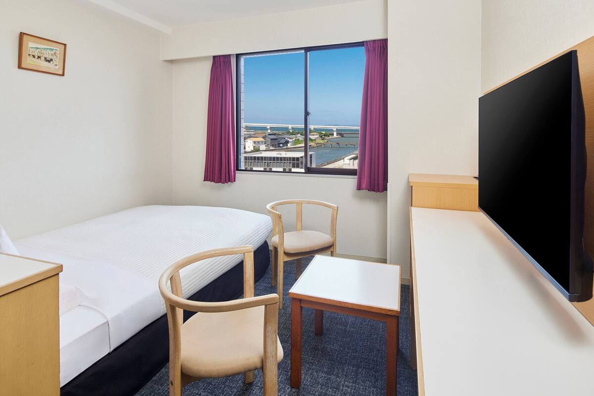 A hotel room with a bed, a desk, a chair, a TV, and a window with a view of a bridge and buildings.
