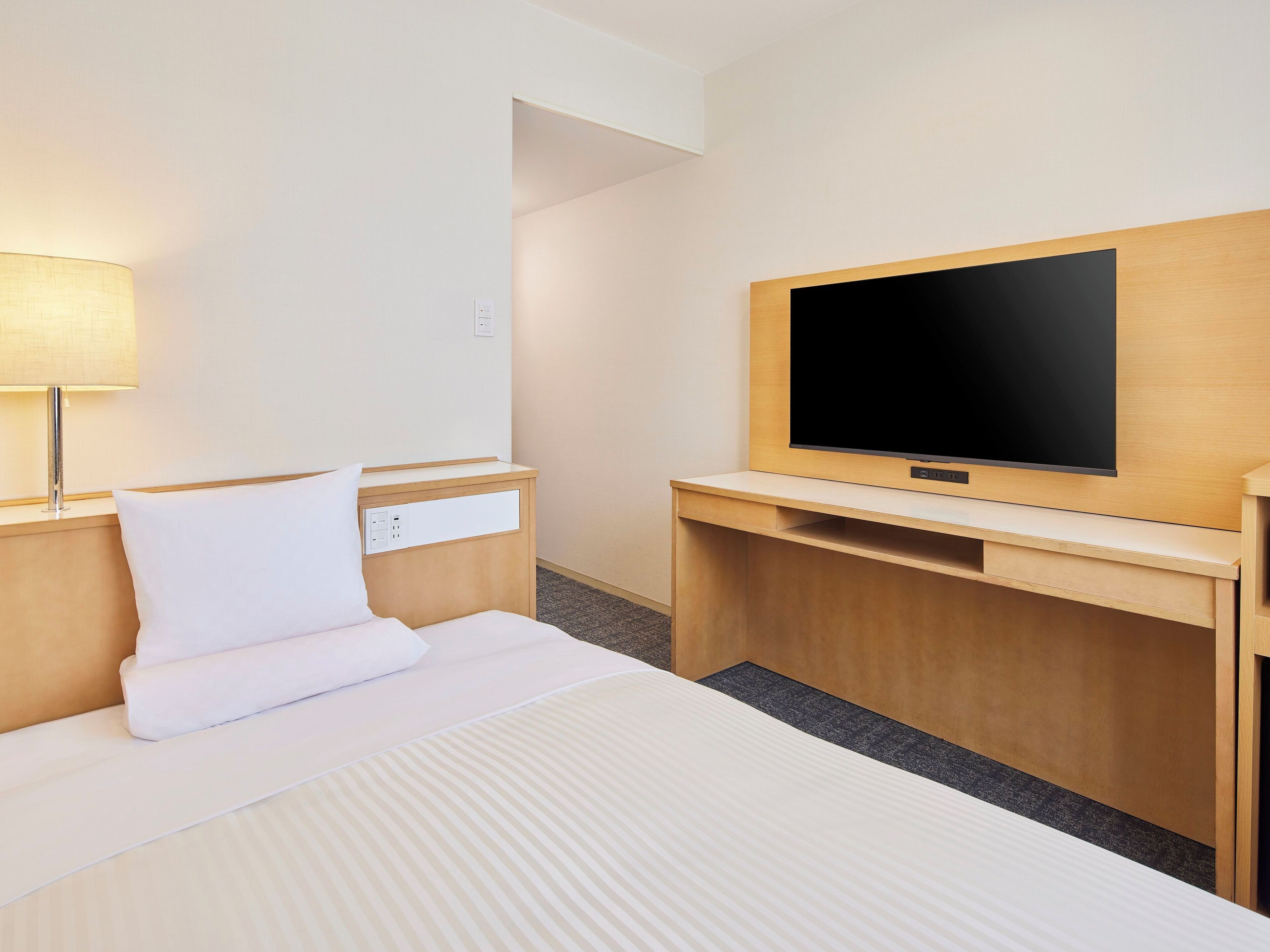 Habitación de hotel con una cama, una mesita de noche con lámpara, un televisor en una pared de madera y un pequeño escritorio con una silla.