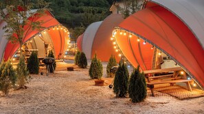 Una hilera de tiendas en forma de cúpula con luces de cadena, una parrilla y pequeños árboles.