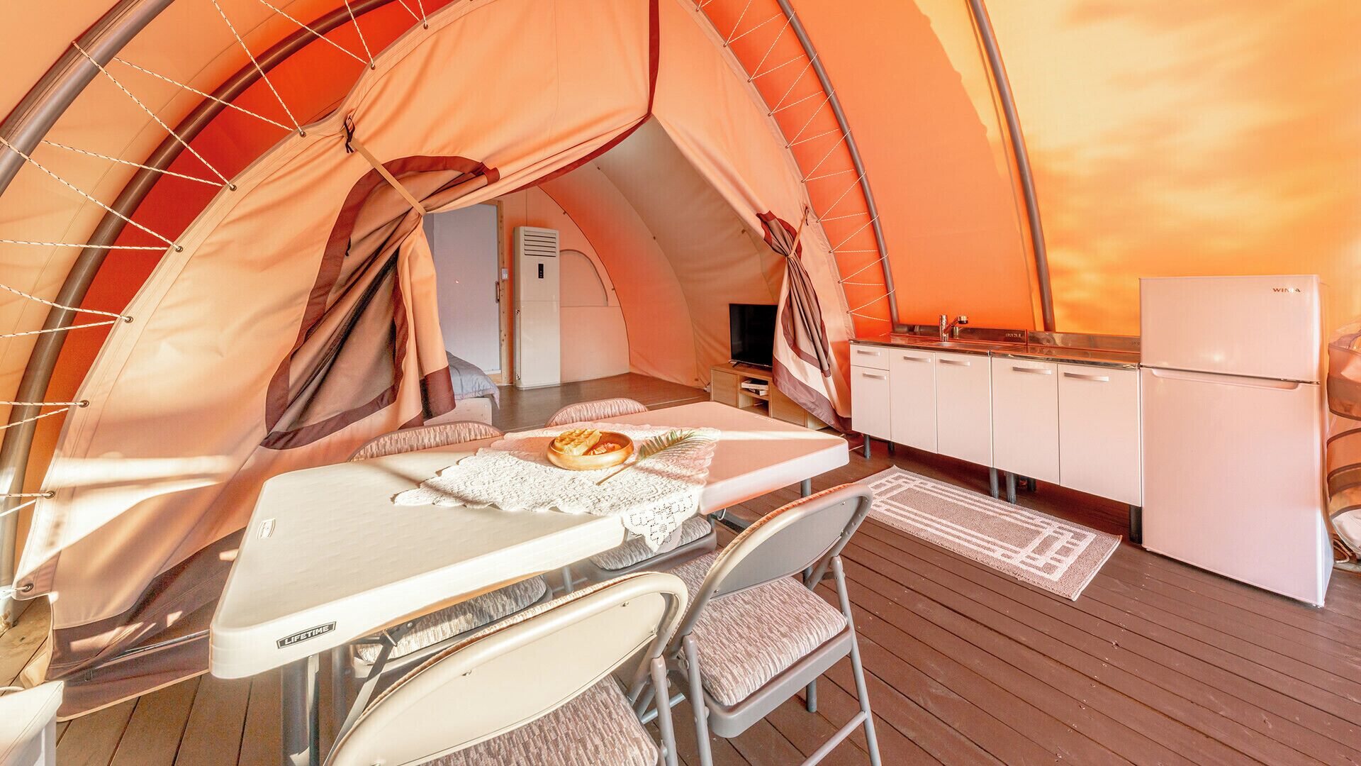 Acolhimento em tenda aconchegante, com área de refeições, cozinha compacta e espaço de estar.