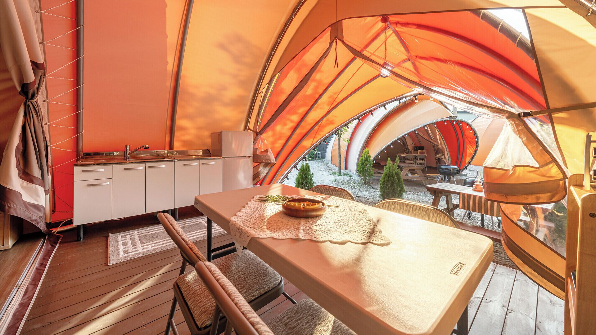 Tienda de campaña con área de cocina, que incluye una mesa, un bol y una silla, y vistas al exterior.