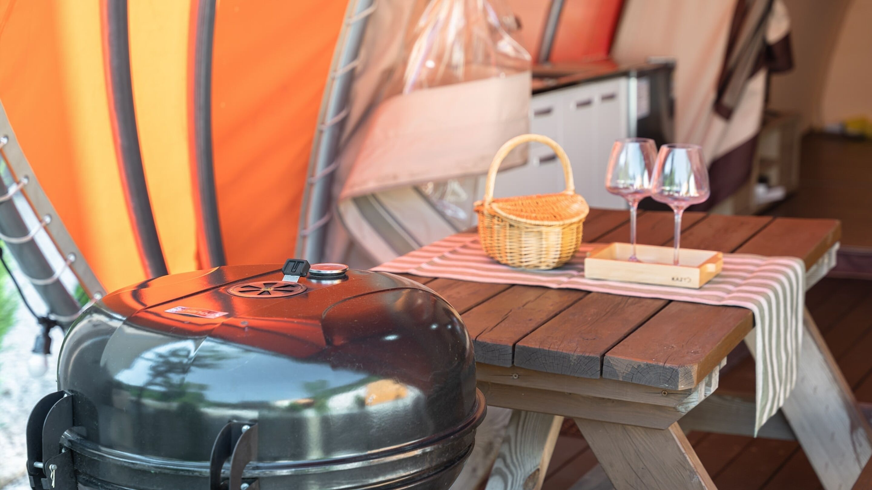 A barbecue grill on a wooden table with a picnic basket and wine glasses.