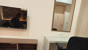 Quarto com televisão presa na parede, uma escrivaninha de madeira, uma cadeira de escritório preta e um cesto de lixo.