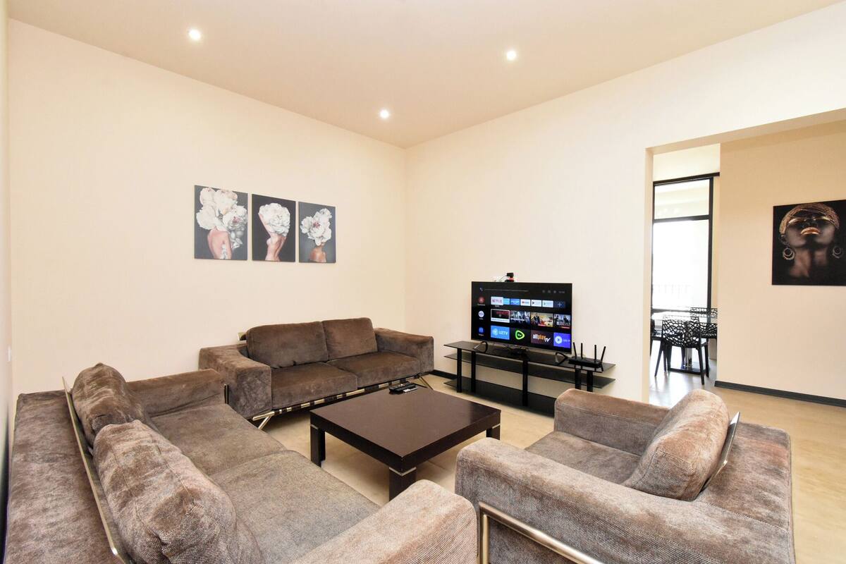 A modern living room with a sectional sofa, a coffee table, and a flat-screen TV. There are wall-mounted artworks and a dining area visible in the background.