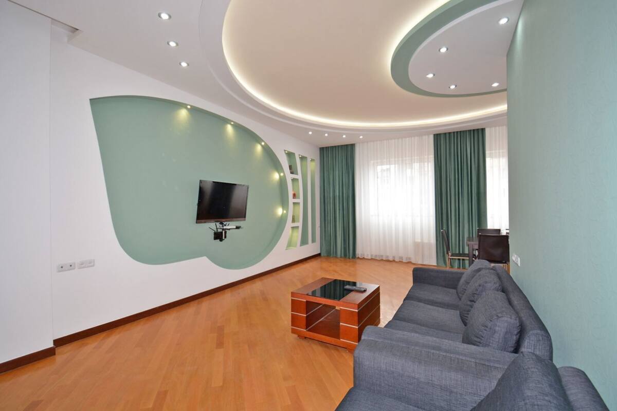 A modern living room with a sectional sofa, a flat-screen TV mounted on a green accent wall, wooden flooring, and a glass coffee table.