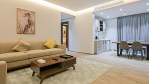A modern living room with a sofa, coffee table, and a painting. There is a dining area with a table and chairs. - Tiara hotel (Jeddah)