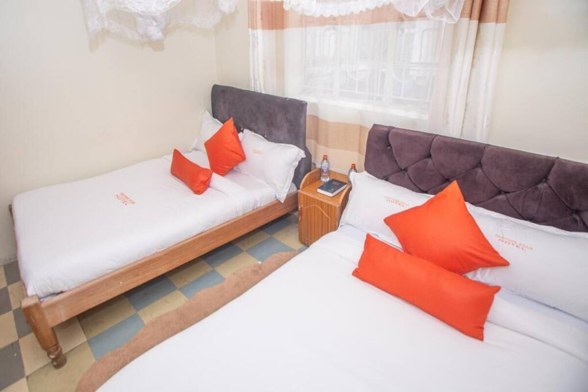 Une chambre d’hôtel avec deux lits simples, chacun doté de draps blancs et d’oreillers orange. On y trouve une petite table de chevet en bois, sur laquelle sont posés une bouteille d’eau et un livre. La chambre a un sol à carreaux et une fenêtre avec des rideaux.