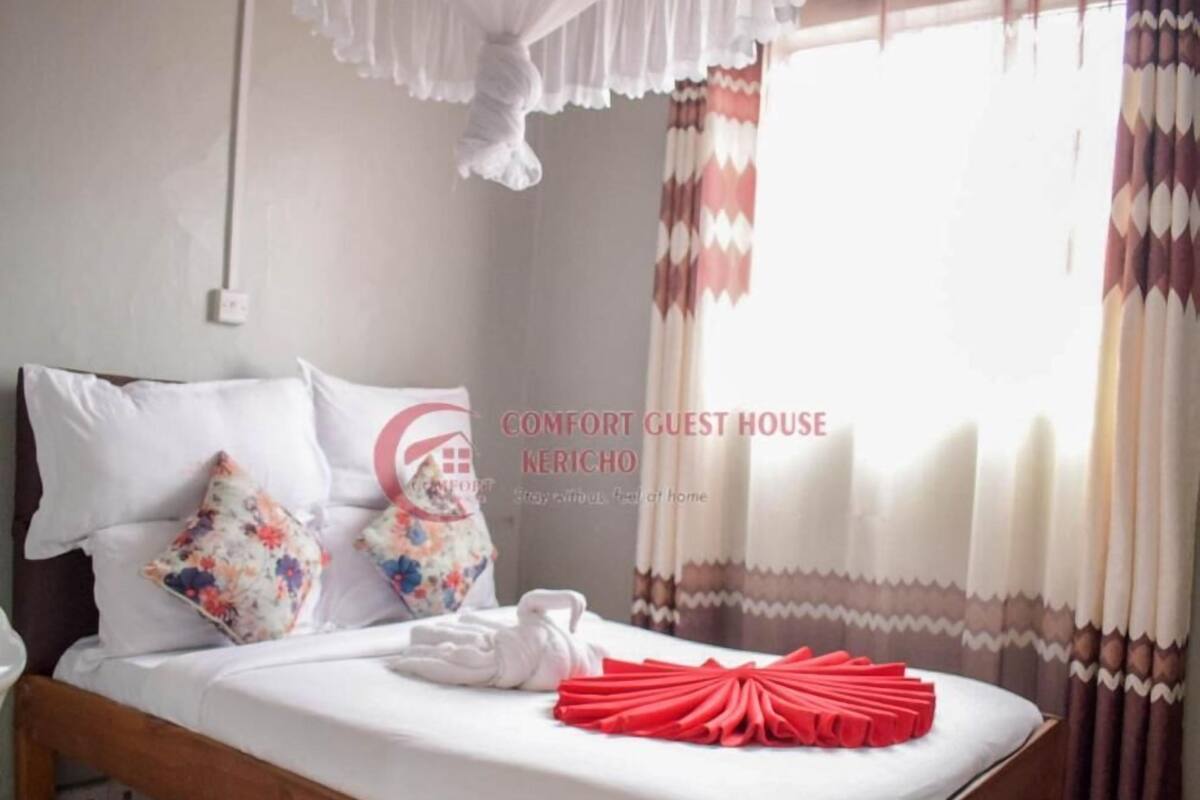 Un lit bien fait, avec des draps blancs, deux coussins à motifs floraux et un objet décoratif rouge posé sur le lit.