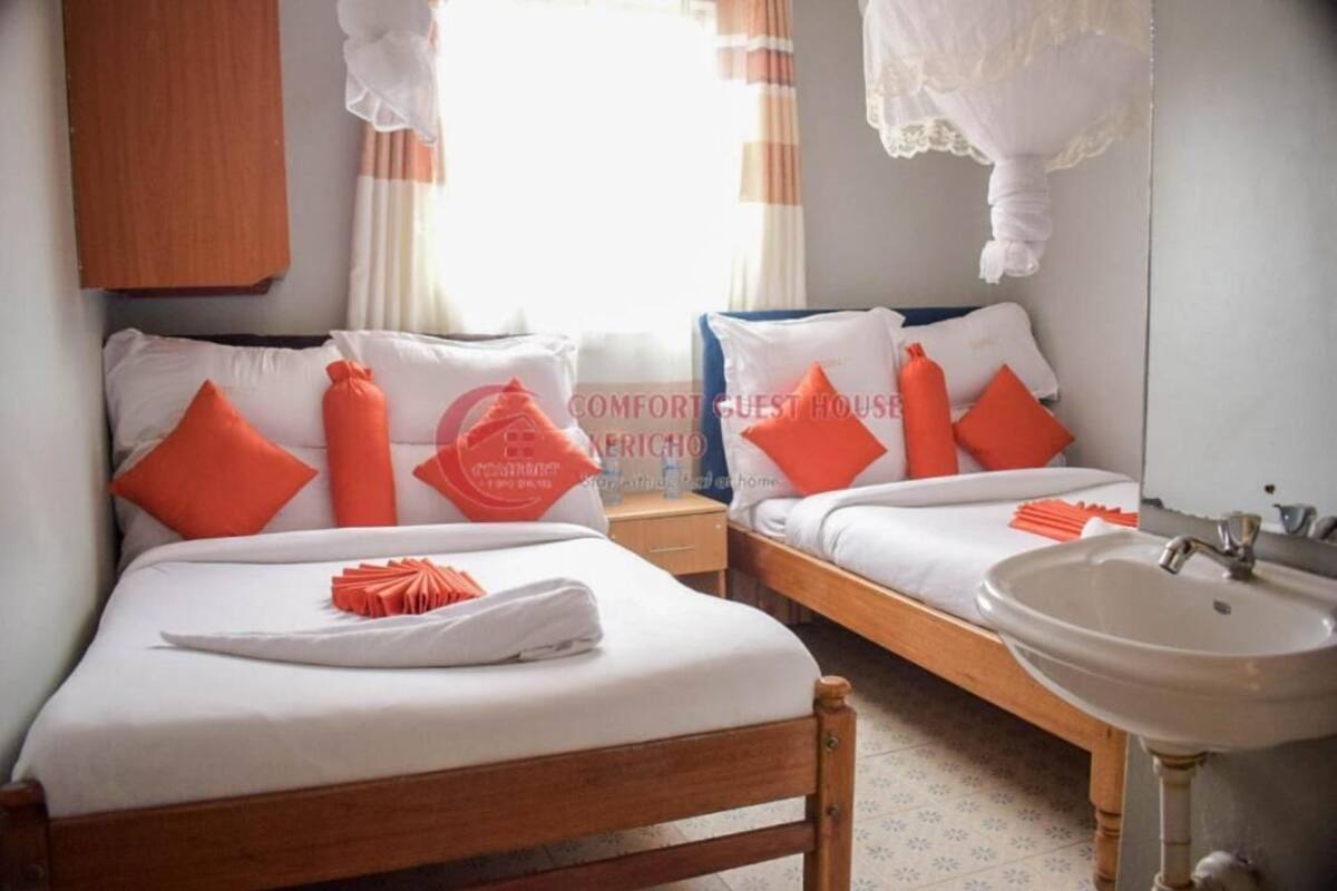 Une chambre d’hôtel avec deux lits, une tête de lit en bois, un lavabo blanc et une fenêtre avec des rideaux.