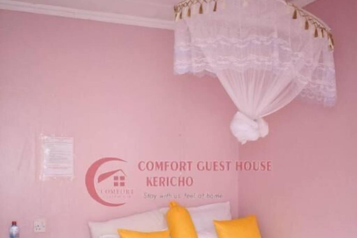 Une chambre d’hôtes aux couleurs rose, avec un lit, des oreillers jaunes, une table de chevet en bois et une chaise.